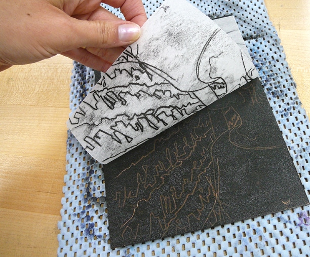 a hand pulling a sheet of paper off a rectangular plate coated with black ink