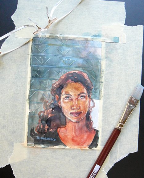 A small watercolor portrait of a woman's face, looking directly at the viewer