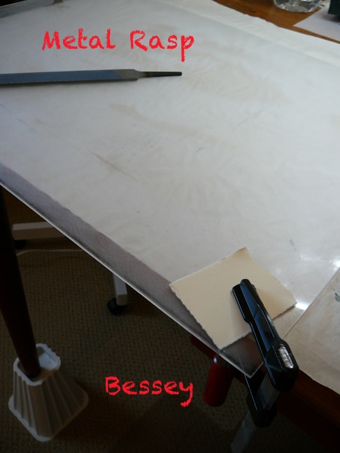 a sheet of polycarbonate clamped to a table being sanded on the surface, and beveled around the edges with a rasp.