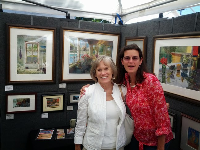 The artist with her arm around a woman standing inside a booth surrounded by paintings at an art festival