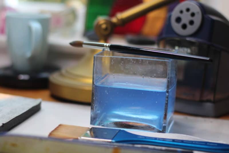 a square glass container with rinse water on an artist's table and a paint brush resting across it's top