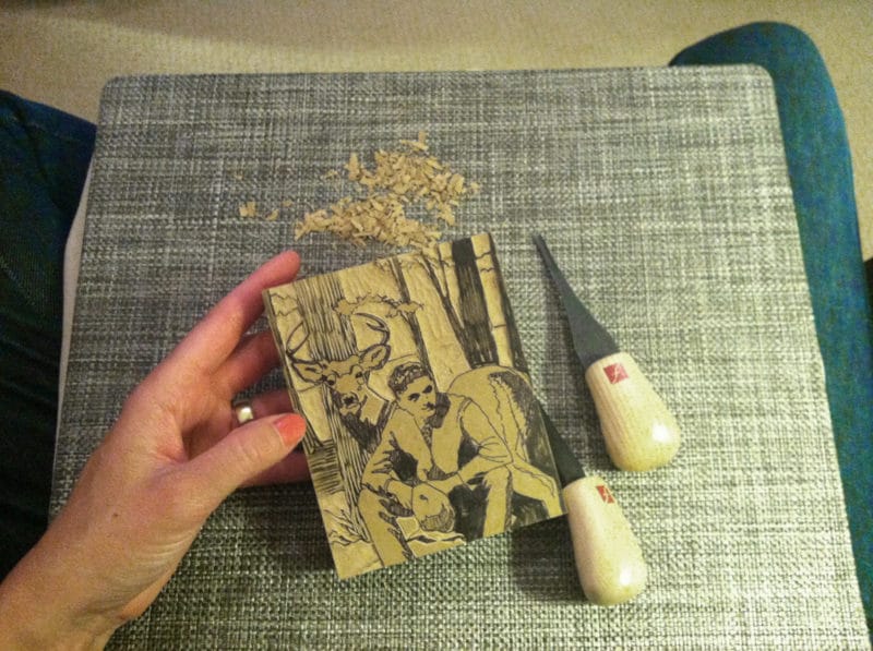 carving a linocut of a man in front of a stag on the couch on a lap desk