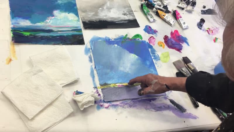 A man's hand scratching into wet paint on a festive, colorful sky-based landscape