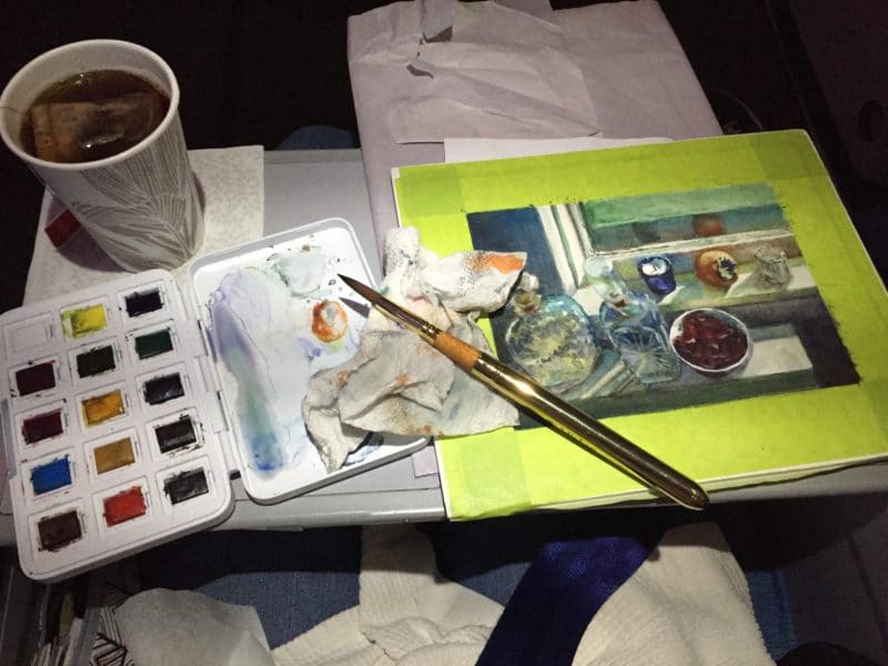 an airplane tray table with a cup of tea, a small travel palette of watercolors and a still life painting in process