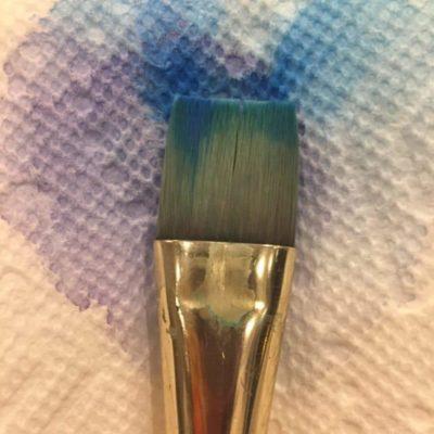a paint brush laying against a apper towel with watercolor that has seeped from the brush and bloomed all over the paper towel in purple and blue