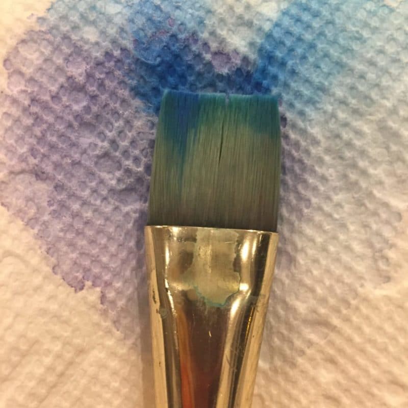 a paint brush laying against a apper towel with watercolor that has seeped from the brush and bloomed all over the paper towel in purple and blue