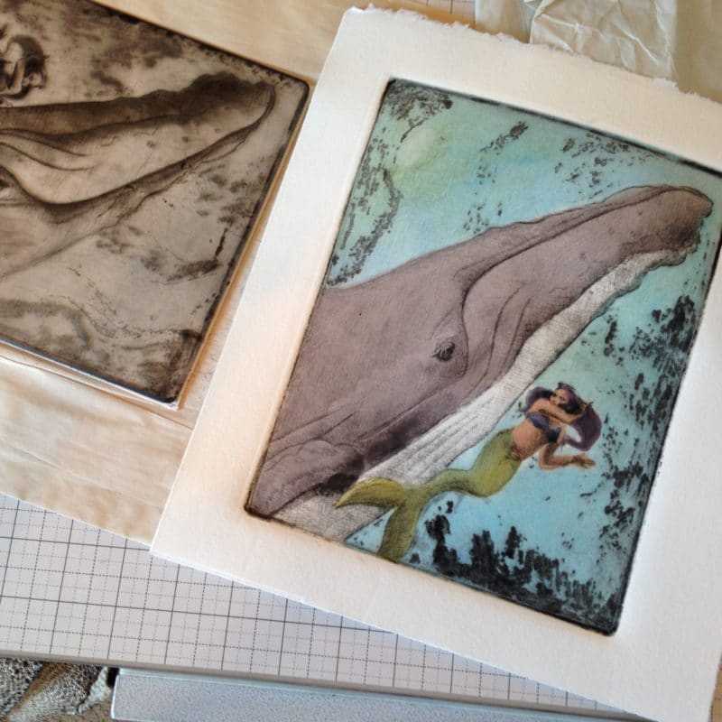 printmaking on a press bed - the intaglio drypoint plate next to the resulting print of a whale and a mermaid