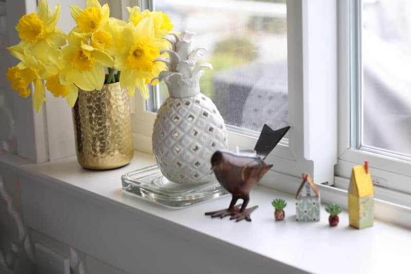 a window sill with yellow daffodils, a ceramic pineapple, a bird candle holder and two little patterned paper houses