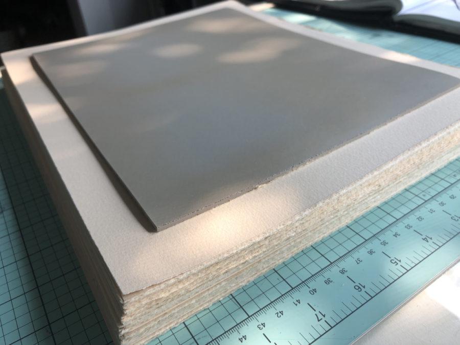 A sanded and prepared sheet of battleship gray linoleum, ready for a design and carving, on a stack of torn-to-size printmaking paper waiting to be editioned.