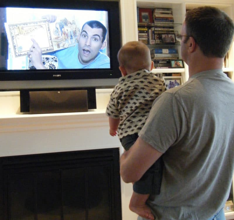 A soldier on a tv screen reading a storybook to an infant held by his grandfather