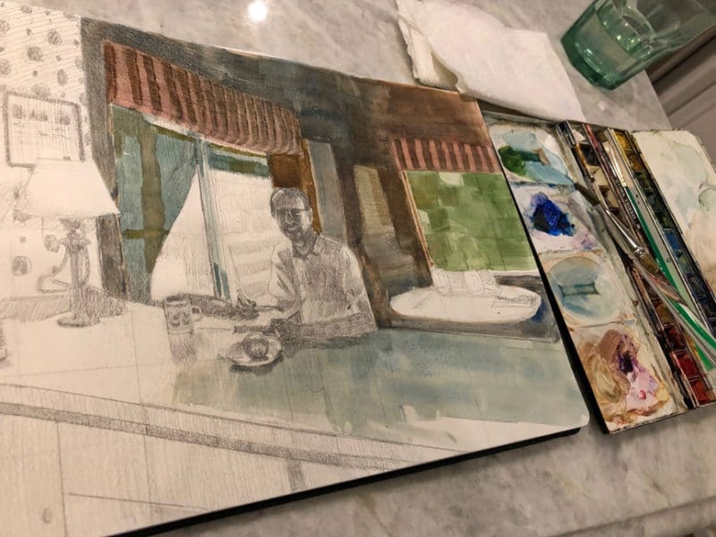 A sketchbook open on a kitchen counter, with watercolor palette and a cup of water, as the first washes of pigments are added to the pencil sketch