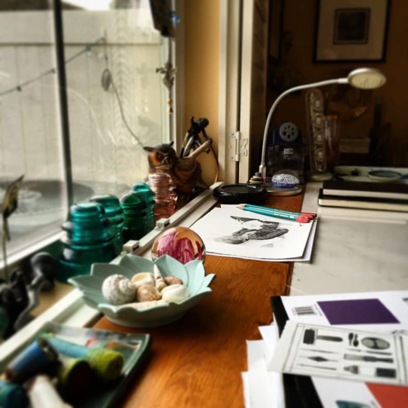 an artist's studio table at a window with printmaking, drawing and painting supplies