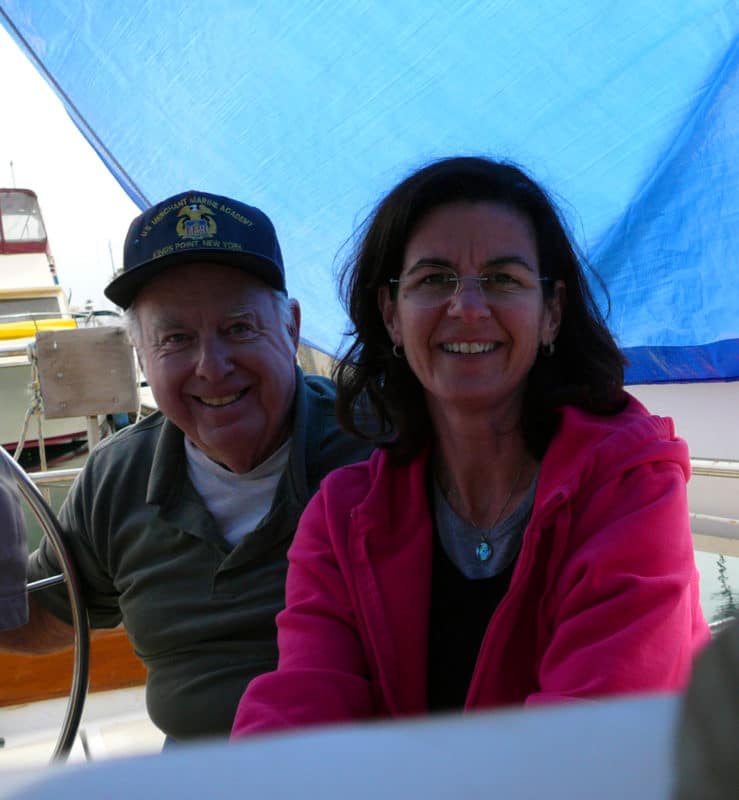 An older man, in a merchant marine cap, and a middle aged woman in a pink hoodie, sitting under a blue tarp in the cockpit of a sailboat.
